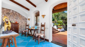 A room with a stone wall, wooden ceiling beams, a tiled floor, a dining table with chairs, and a view of a garden through an open doorway. - YourHome - Villa Mia (Massa Lubrense)