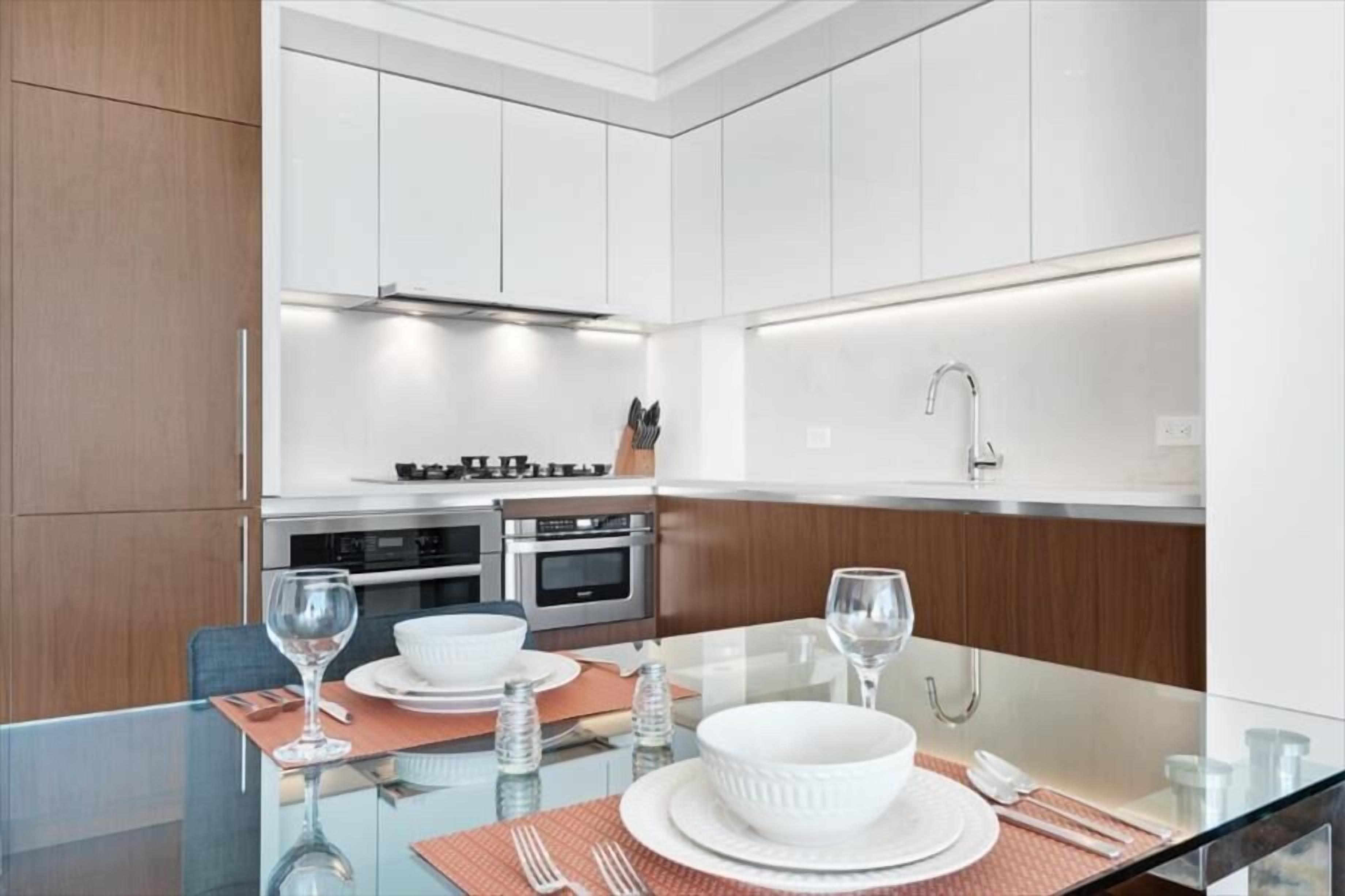A modern kitchen with white cabinets, stainless steel appliances, and a glass dining table set for two.
