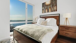 Un dormitorio con un ventanal grande, una cama con ropa de cama blanca, un cabecero de madera y un cuadro en la pared.