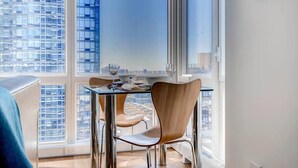 Un espace de repas moderne avec une table et des chaises, de grandes fenêtres offrant une vue sur la ville et un immeuble de grande hauteur en arrière-plan.