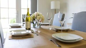 Una mesa de comedor dispuesta con platos, cubiertos, una botella de vino y copas, con un jarrón de flores y una lámpara al fondo.