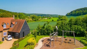 Una casa de campo con entrada para vehículos, un automóvil y un parque infantil con columpios y estructuras para escalar.