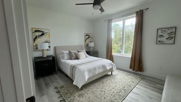 Un dormitorio con cama, mesitas de noche, ventilador de techo, ventana con cortinas y obras de arte en las paredes.