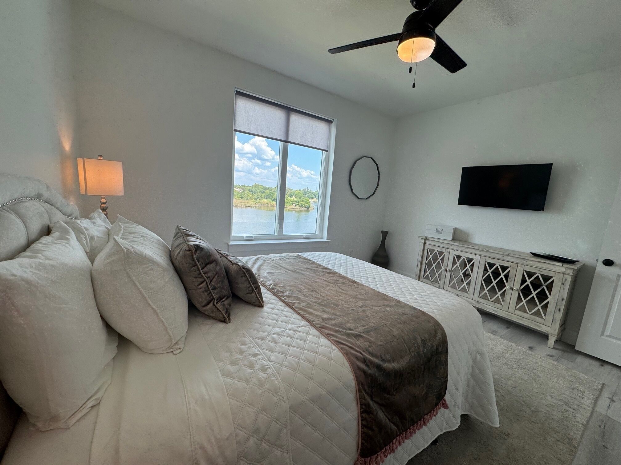 A bedroom with a bed, a window with a view, a ceiling fan, a wall-mounted TV, and a radiator.