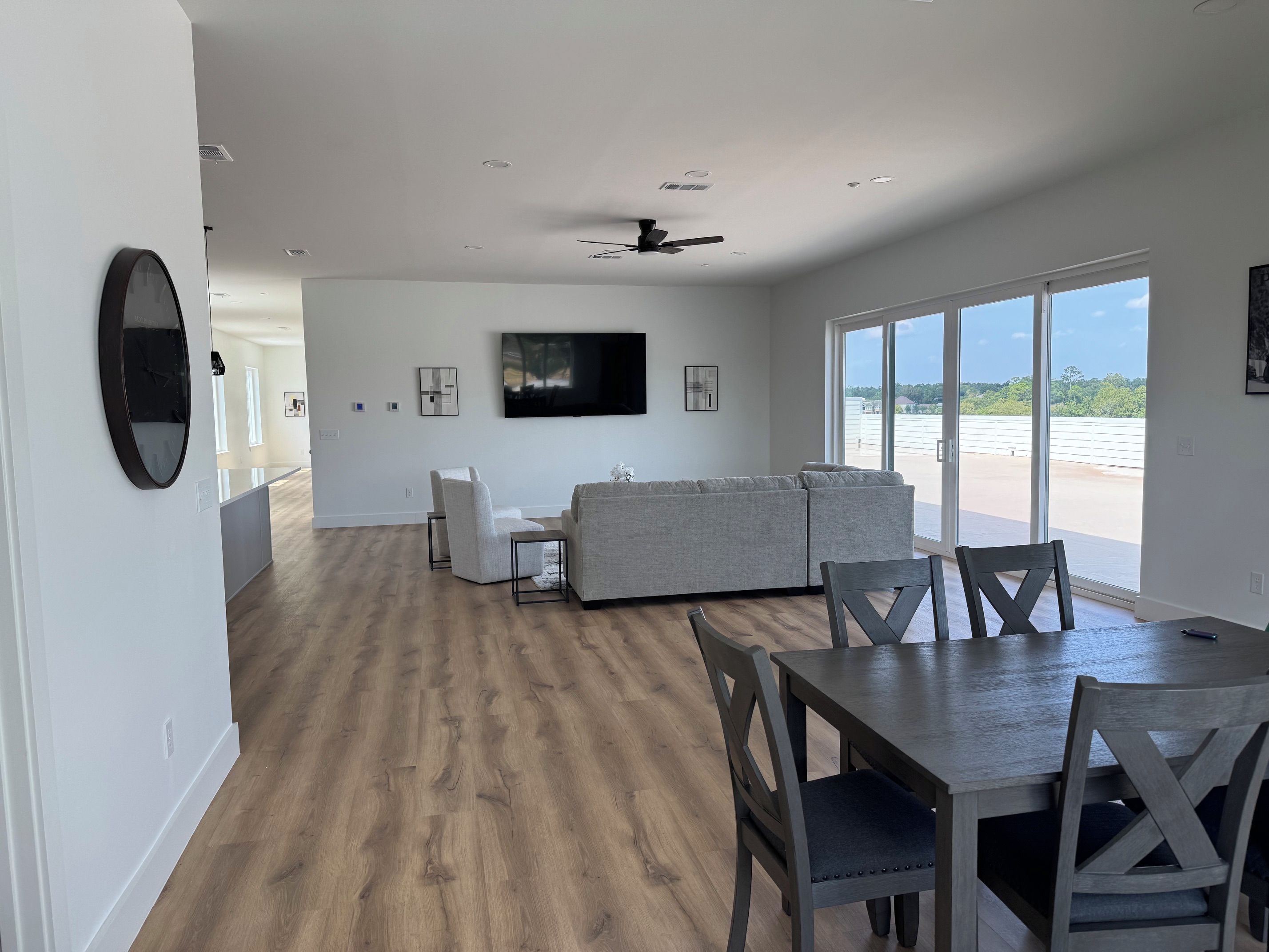 A spacious living area with a large window, a gray sofa, a dining table with chairs, and a flat-screen TV mounted on the wall.