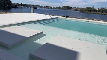 A swimming pool with clear water, surrounded by white steps and a view of a river and distant houses.