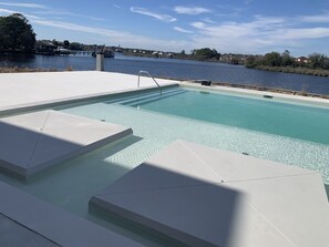 A swimming pool with clear water, surrounded by white steps and a view of a river and distant houses. - Handsboro Pointe Condo & Suites (Gulfport)