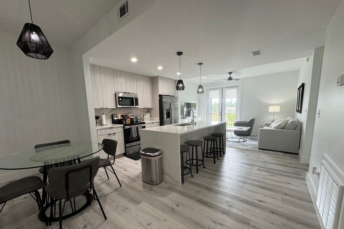 A modern kitchen with a central island, bar stools, a dining area with a glass table, and a living room with a sofa and a lamp.