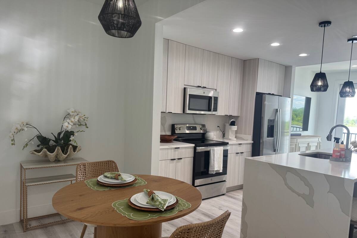 A modern kitchen with white cabinetry, stainless steel appliances, and a dining area with a wooden table and wicker chairs.