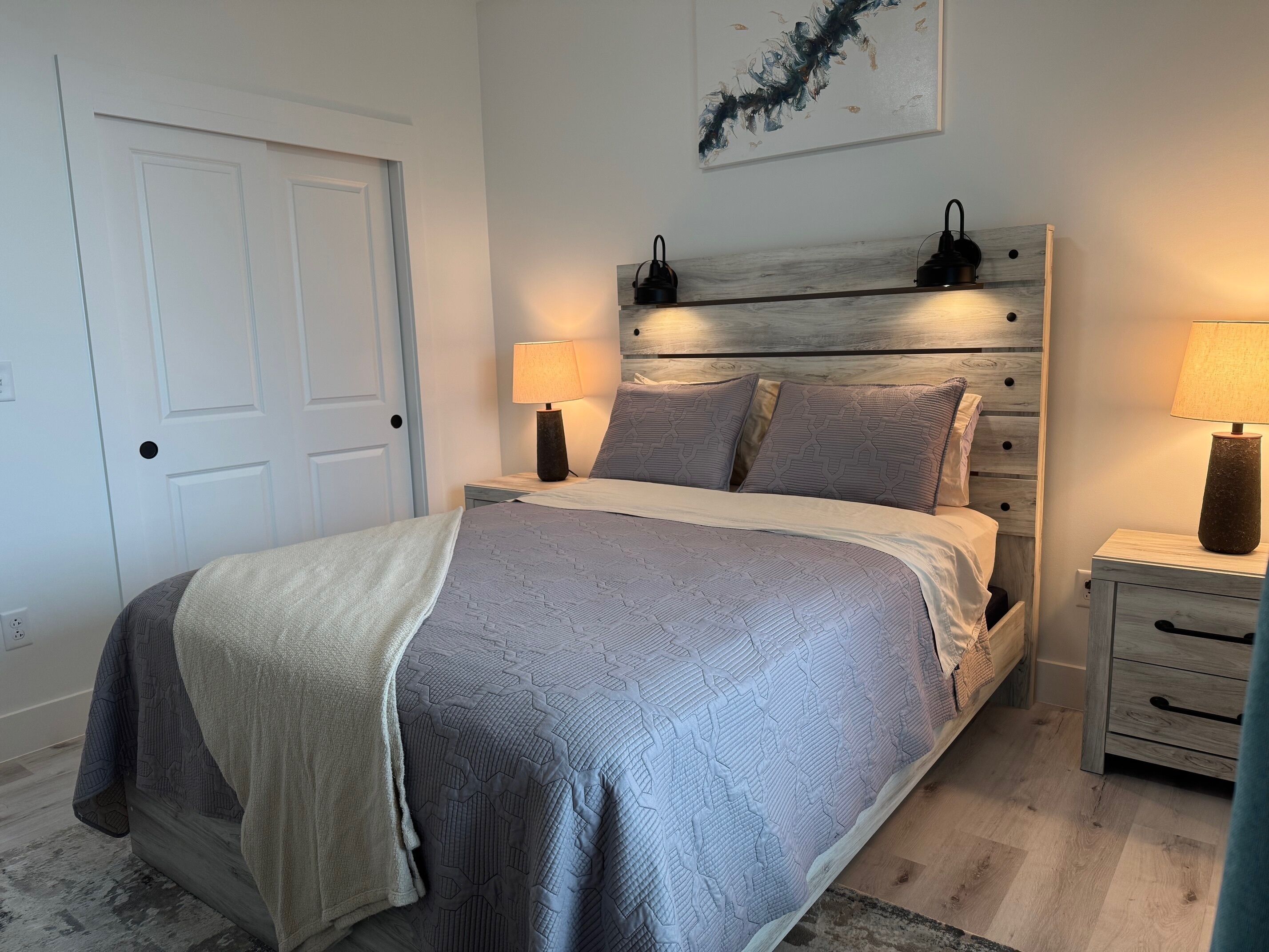 A bedroom with a bed, two bedside tables, a door, and a wall-mounted artwork.
