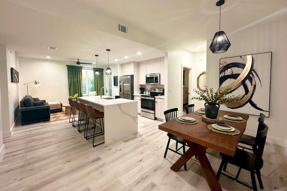 A modern kitchen with a central island, dining table with chairs, and a large abstract painting on the wall.