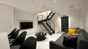 Un salon moderne avec un escalier en colimaçon noir, un téléviseur à écran plat fixé au mur, un canapé gris agrémenté d’un coussin jaune et une table basse noire.