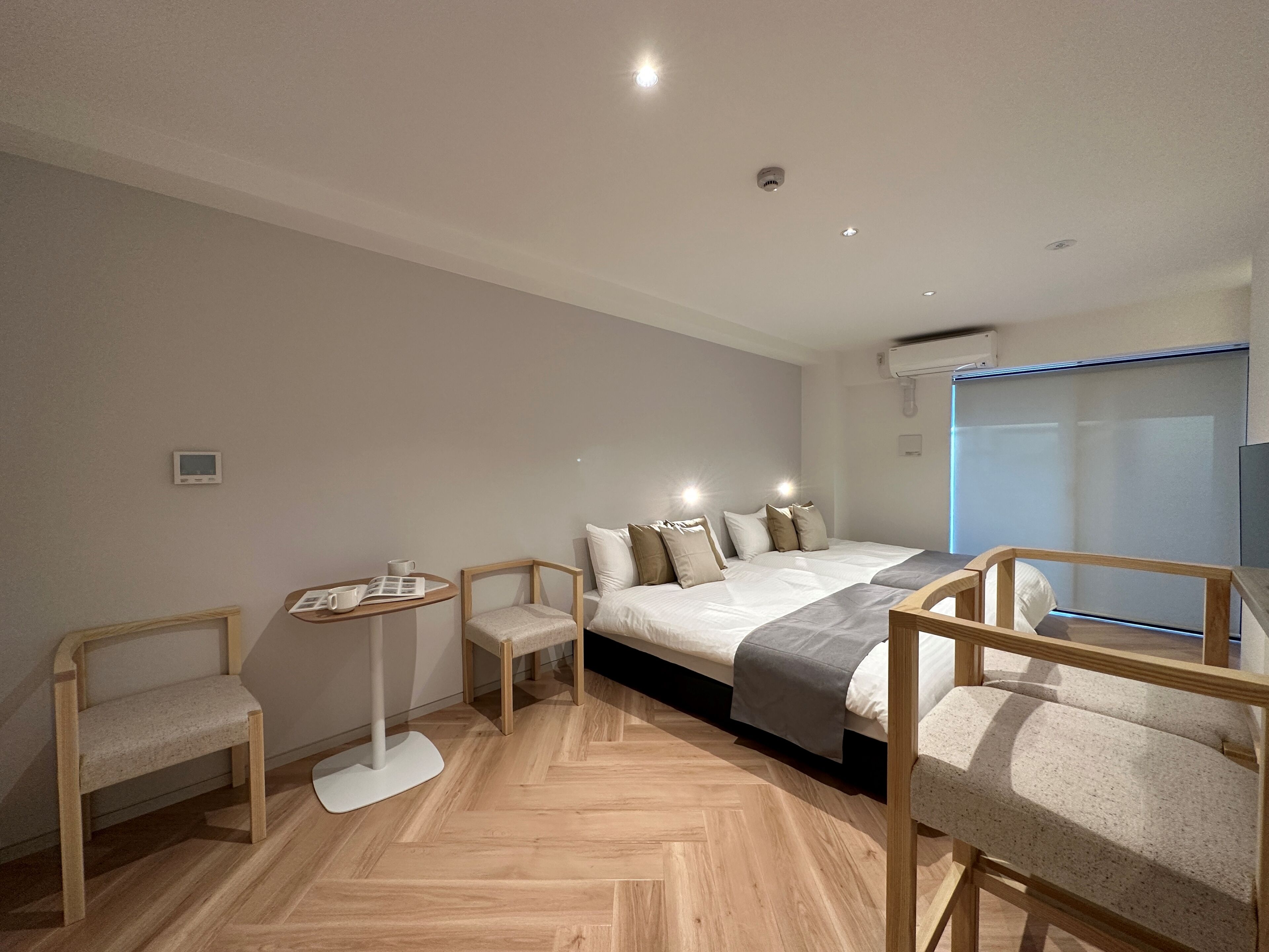 A modern hotel room with a large bed, a small table, two chairs, and a window with blinds.