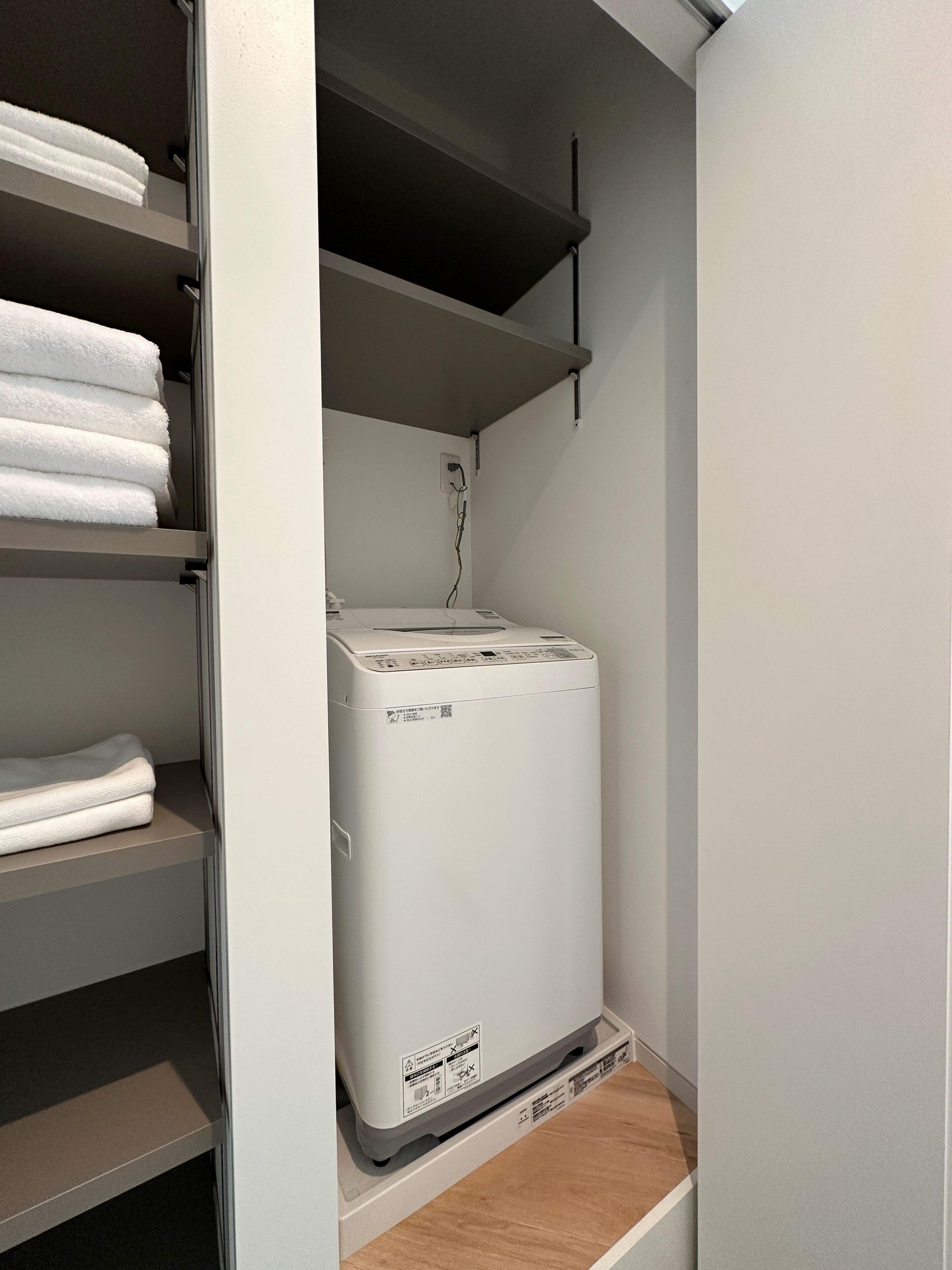 A compact, white laundry machine in a small, enclosed laundry room with shelves above holding folded towels.