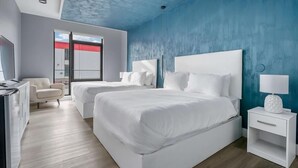 Moderni hotellihuone, jossa on kaksi valkoista sänkyä, työpöytä, tuoli ja näkymä ulos.