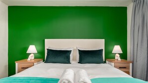 Un lit bien fait, avec du linge de lit blanc et deux serviettes enroulées, un mur vert et deux tables de chevet avec des lampes.