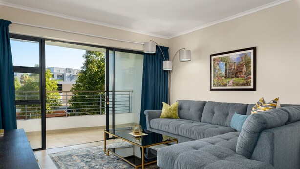 Un salon moderne avec un canapé gris, une table basse en verre et une vue sur un balcon agrémenté d’un rideau bleu.