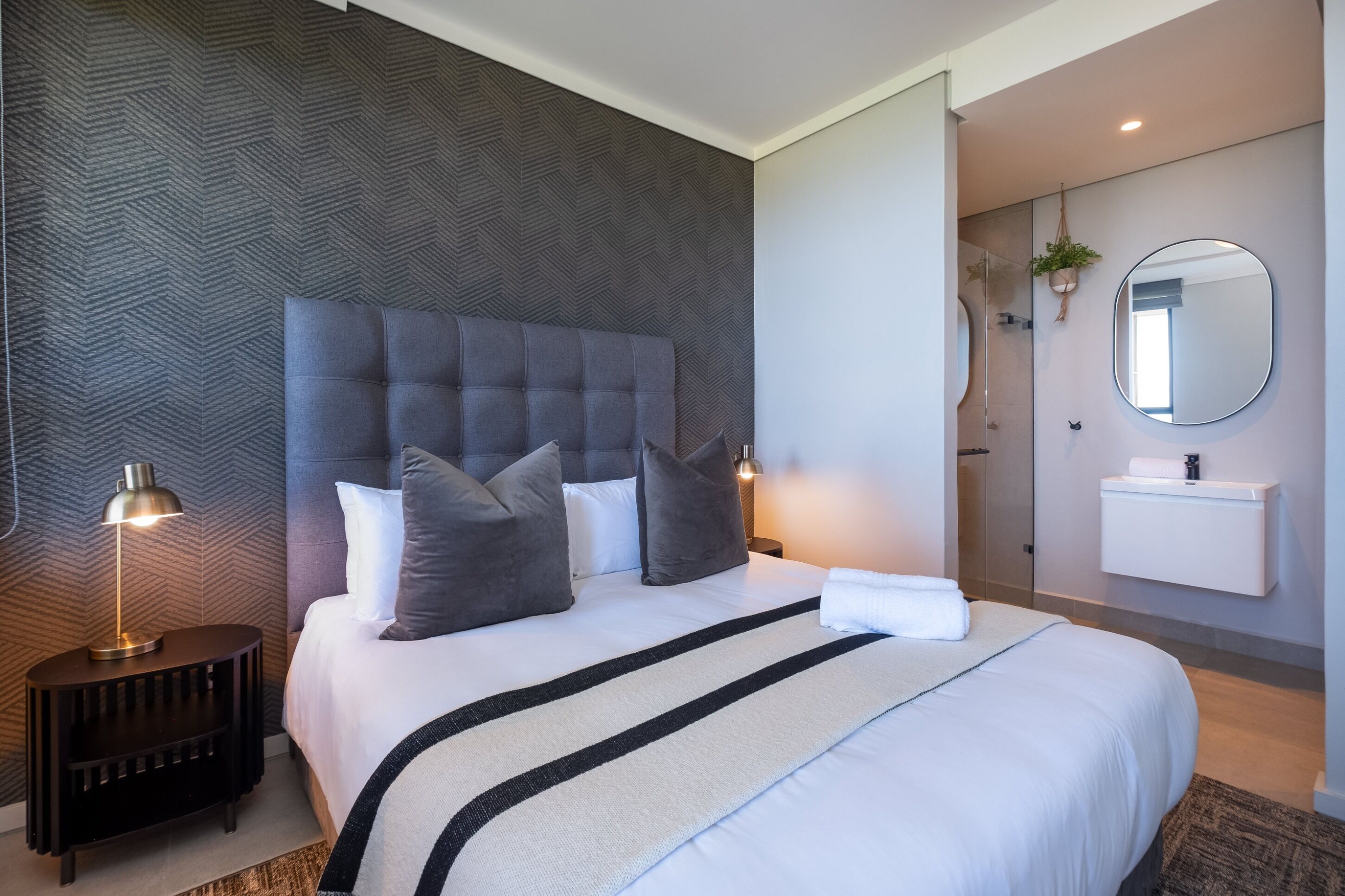 Ein modernes Hotelzimmer mit einem großen Bett, einem Nachttisch mit Lampe, einem runden Spiegel und einem kleinen Topfpflanzen.