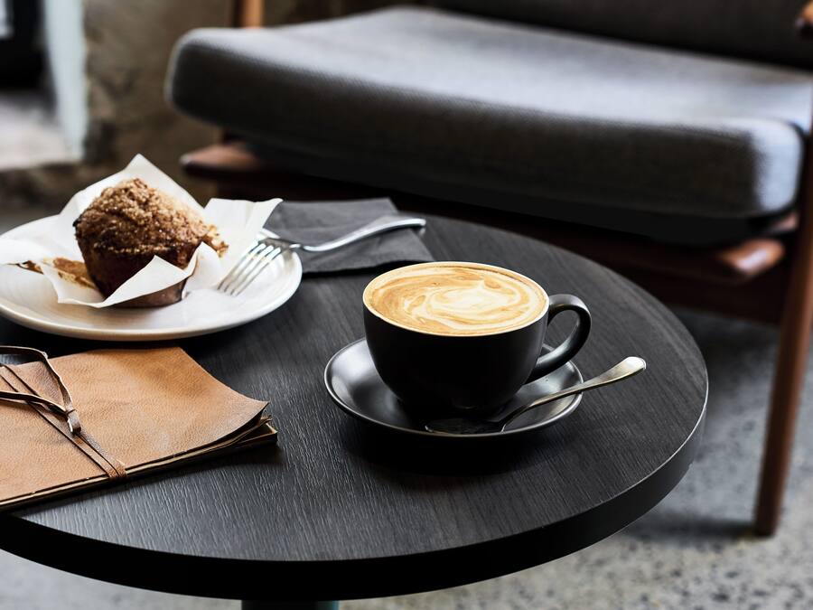 Eine schwarze Tasse Kaffee mit Latte-Art auf einem schwarzen Untertasse, ein Muffin auf einem weißen Teller, eine braune Ledertasche und ein schwarzer Stuhl im Hintergrund.