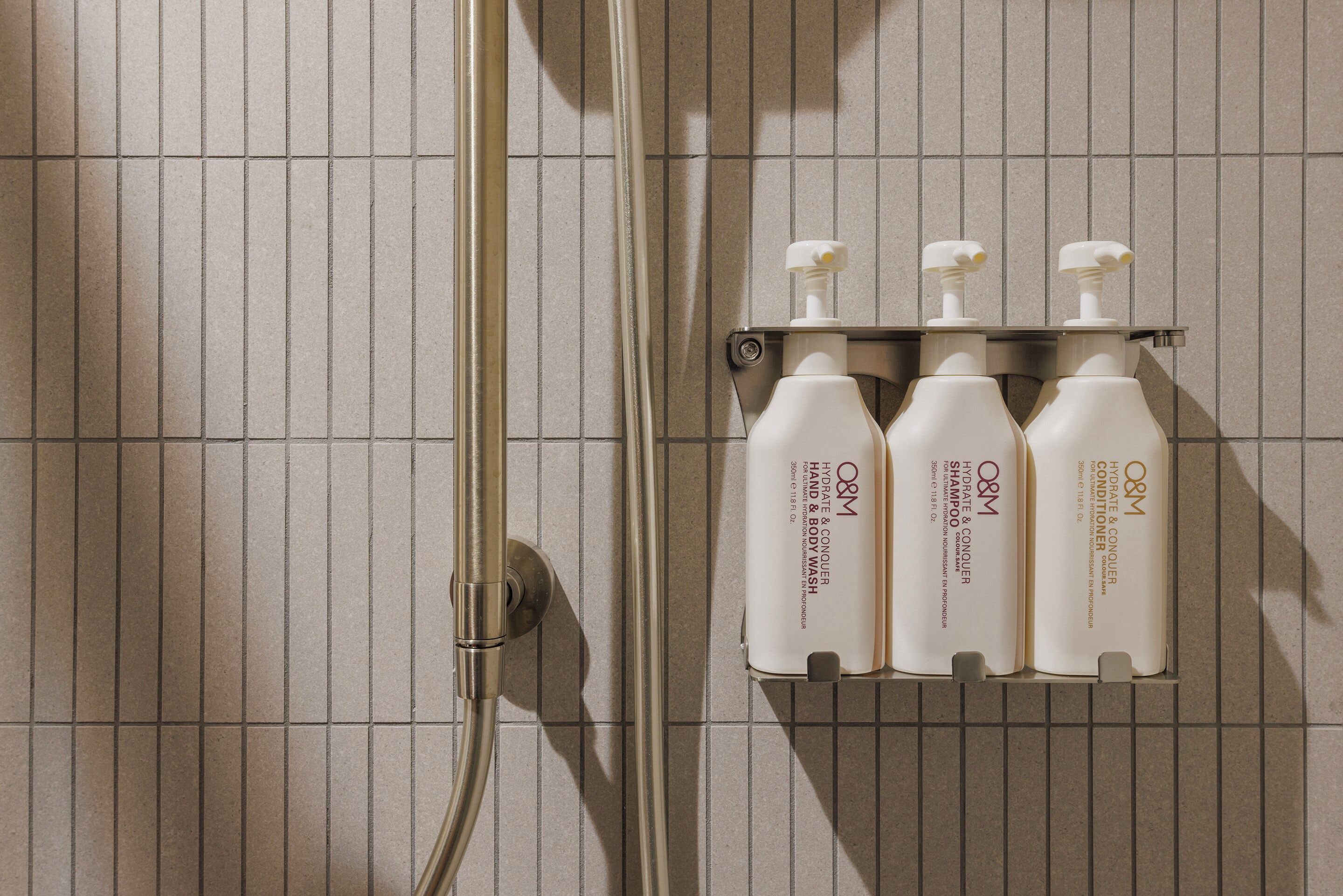 A bathroom with a tiled wall, a shower, and a shelf holding four bottles of O&M hand and body wash.