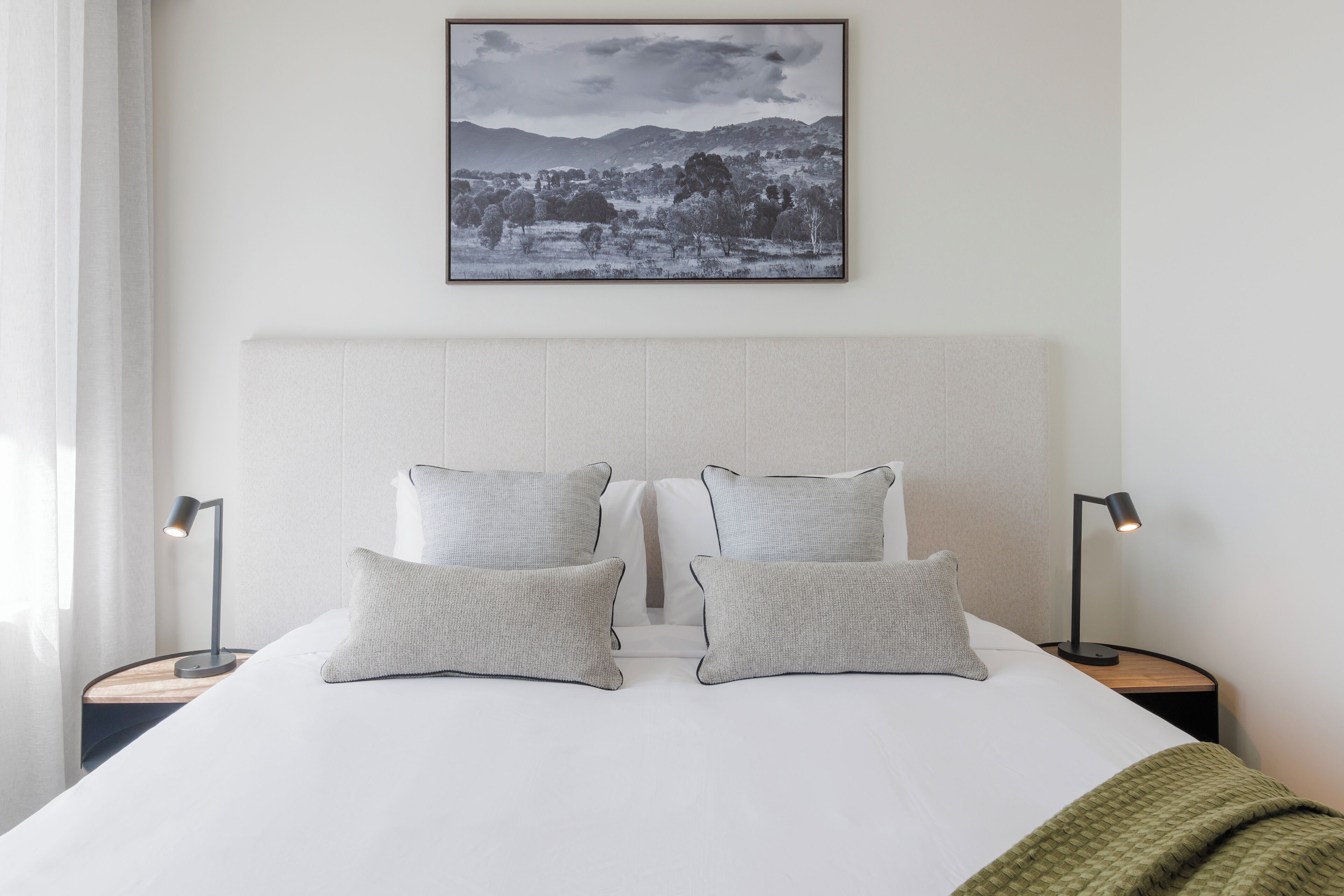 Ein ordentlich bezogenes Bett mit zwei Kissen und zwei Nachttischlampen, ein gerahmtes Landschaftsfoto über dem Bett und eine grüne Decke am Fußende.