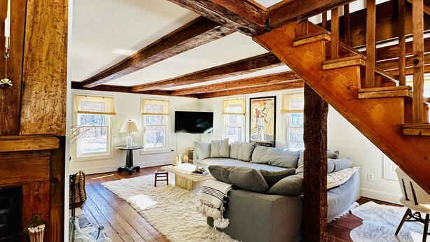 Un loft spacieux avec des poutres apparentes, un coin salon confortable avec un canapé gris, une cheminée et un escalier en bois.