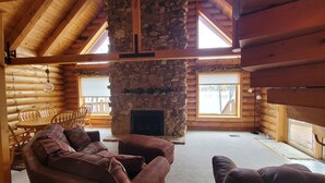 Interior de una cabaña de madera con chimenea de piedra, muebles de madera y ventanas grandes.