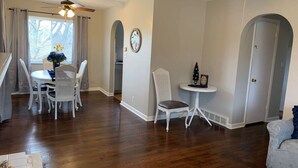 A spacious living room with hardwood flooring, a dining area with white chairs, a small table, and a ceiling fan.