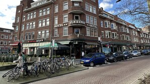 Un hotel de varios pisos con un llamativo letrero "FLEX-INN", fachada de ladrillo y una fila de coches y bicicletas estacionados al frente.