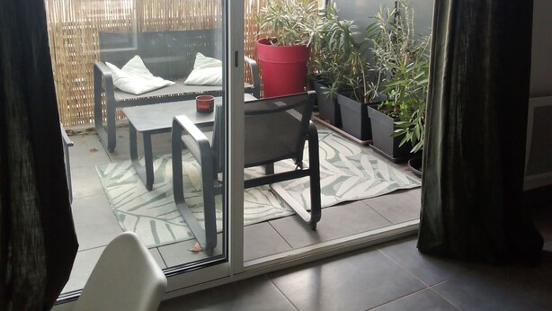 Una habitación con un ventanal corredero que da a un balcón. El balcón tiene una mesa, sillas y plantas en macetas.
