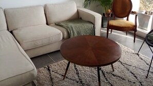 Una sala de estar con un sofá color beige, una silla de madera, una mesa redonda de centro, un tapete estampado y dos cuadros en la pared.
