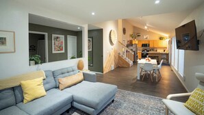 A modern living room with a grey sectional sofa, a dining area with a white table and chairs, and a kitchen in the background.