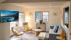 A living room with a large TV, a sofa, a chair, a coffee table, and a window with a view of the beach.