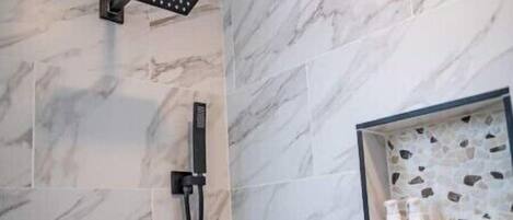 Un baño moderno con ducha, azulejos de mármol, un estante empotrado con artículos de aseo y un panel de control.
