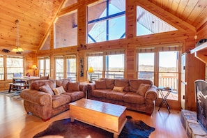 A spacious wooden cabin with large windows, a leather sofa set, a wooden coffee table, and a stone fireplace.