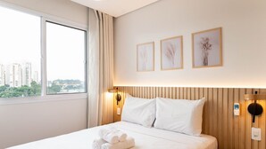 A hotel room with a large bed, a bedside table, a nightstand, and a window with a view of buildings. There are three framed pictures on the wall above the bed. - Studio Laguna (São Paulo)