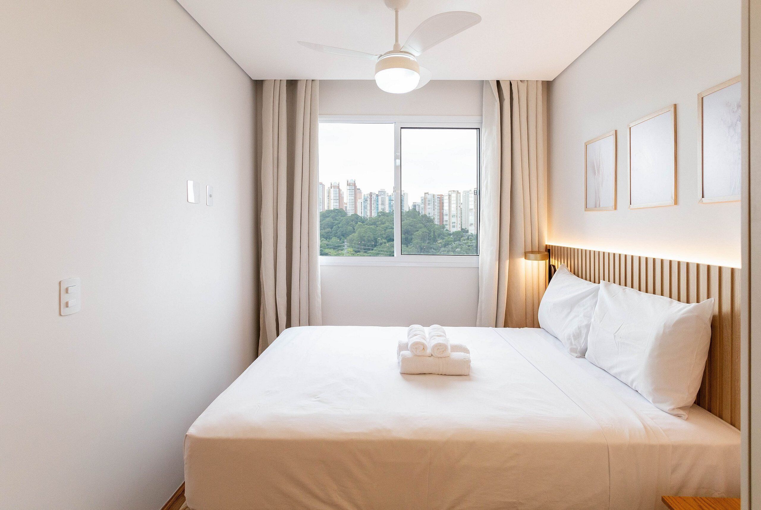 Hotellihuone, jossa on suuri sänky, kattotuuletin, ikkuna verhoilla ja seinällä tauluja.