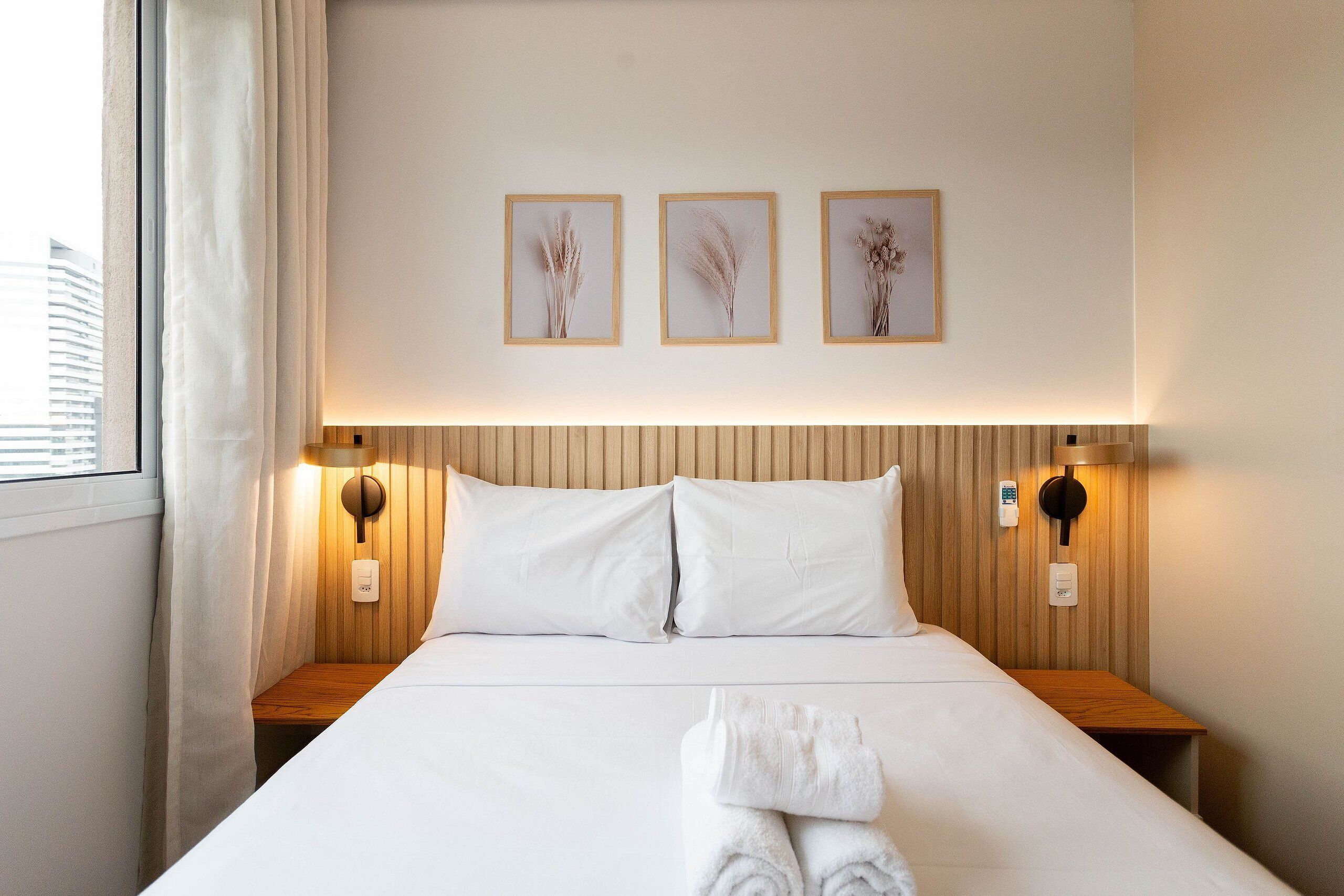 Une chambre d’hôtel avec un grand lit, deux tables de chevet, une lampe fixée au mur et trois tableaux encadrés au-dessus du lit.