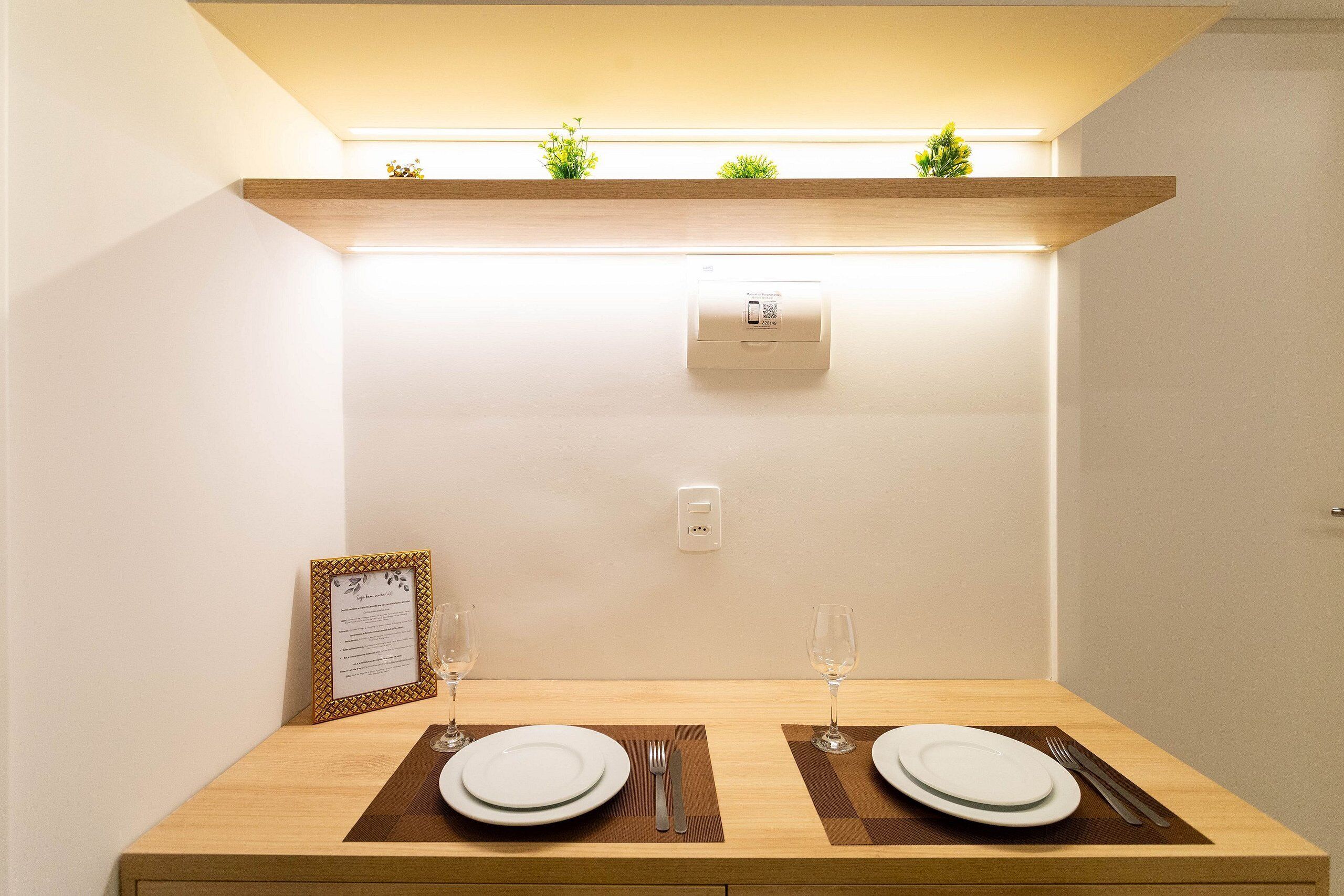 A neatly set dining table with two plates, cutlery, and glasses, a framed menu, and a small plant on a shelf above.