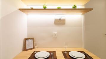A neatly set dining table with two plates, cutlery, and glasses, a framed menu, and a small plant on a shelf above.