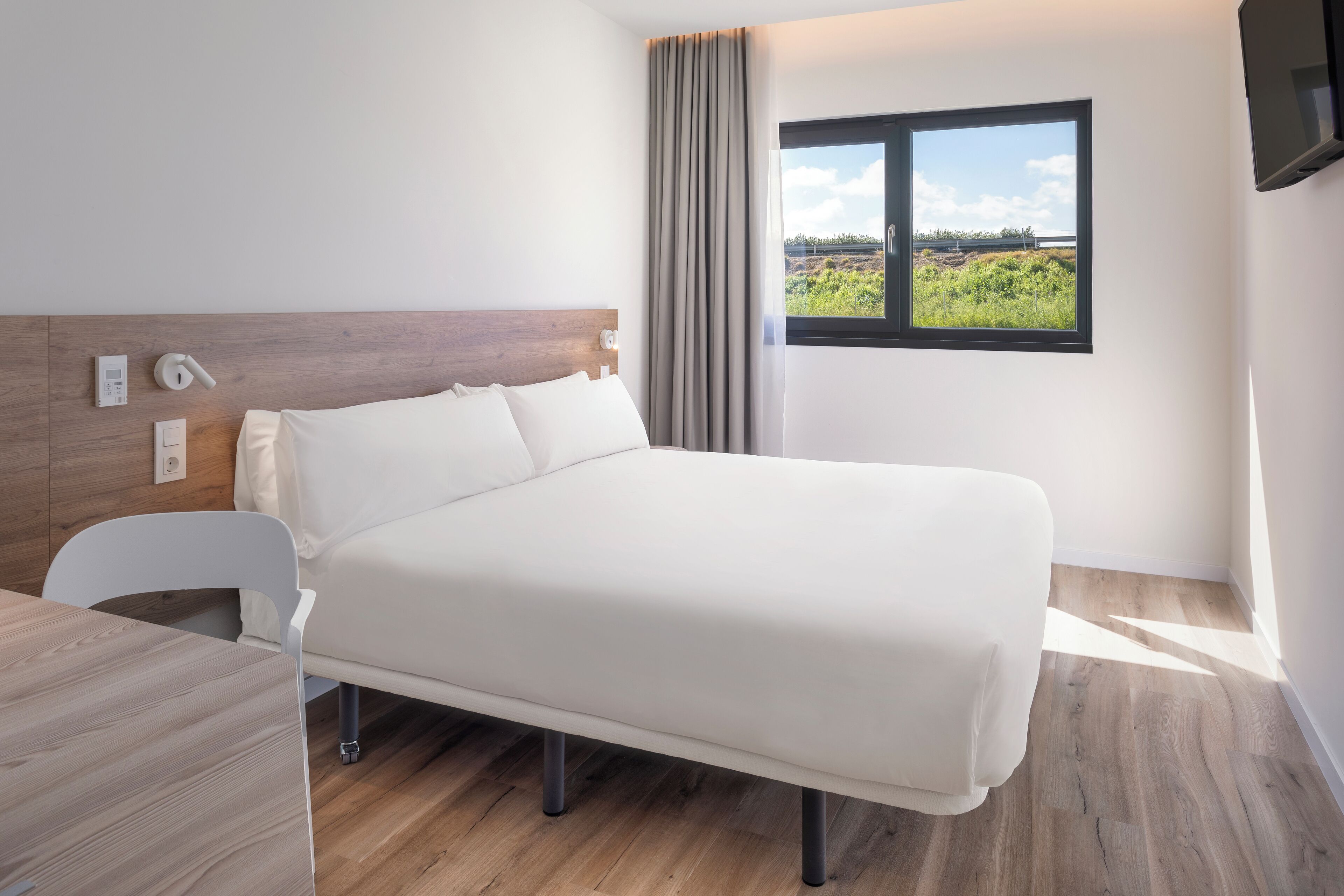 A modern hotel room with a wooden headboard, a bed with white linens, a wooden desk, a chair, a window with a view of greenery and a bridge, and a flat-screen TV mounted on the wall.