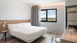 A hotel room with a bed, bedside table, chair, and a window with a view of the city.