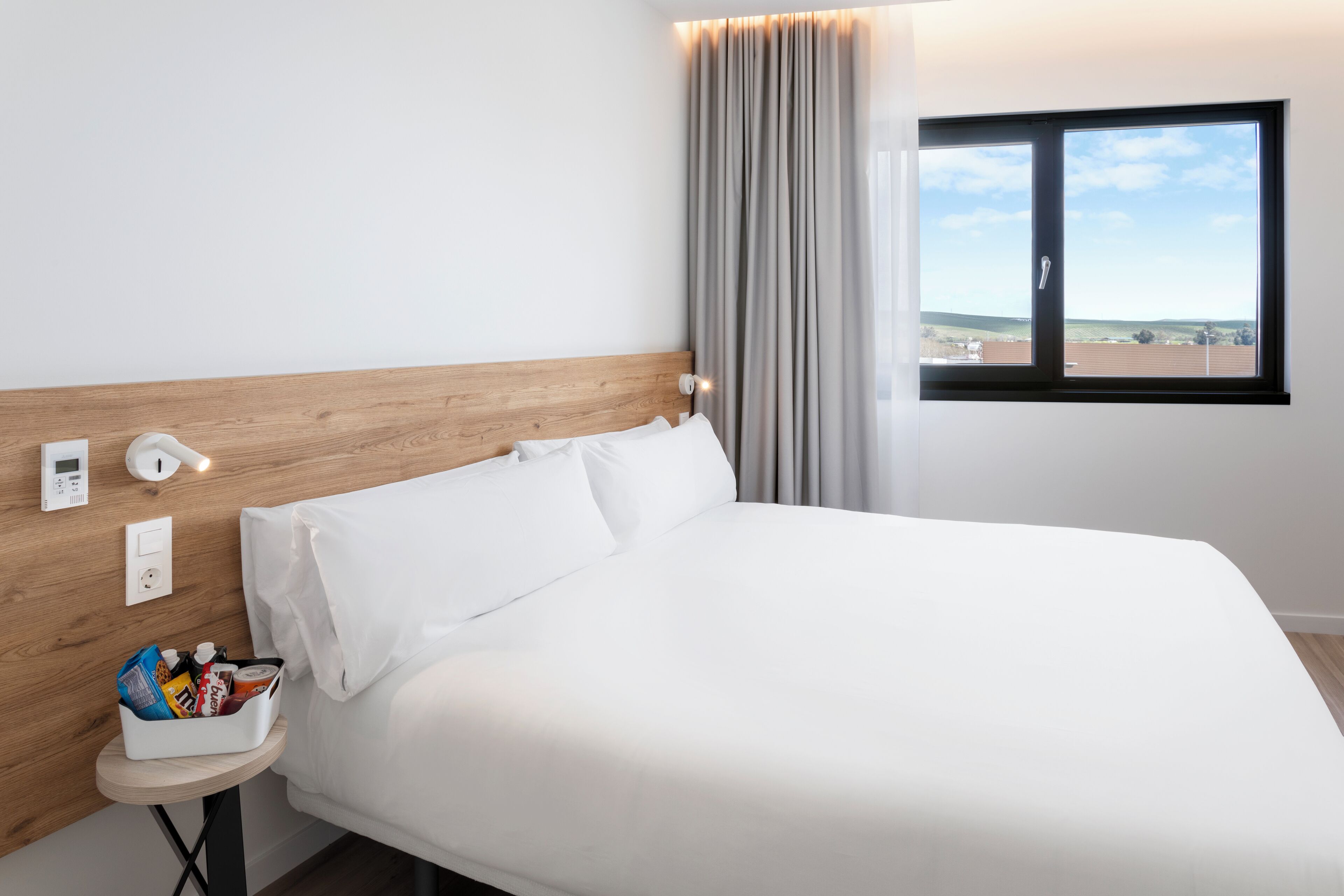 A neatly made bed in a hotel room with a wooden headboard, a bedside table with a snack basket, a window with a view of the landscape, and a control panel on the wall.