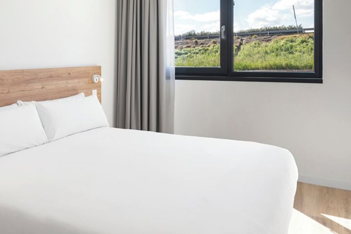 Une chambre d’hôtel moderne avec une tête de lit en bois, un lit blanc, une table de chevet et une fenêtre donnant sur un paysage verdoyant.