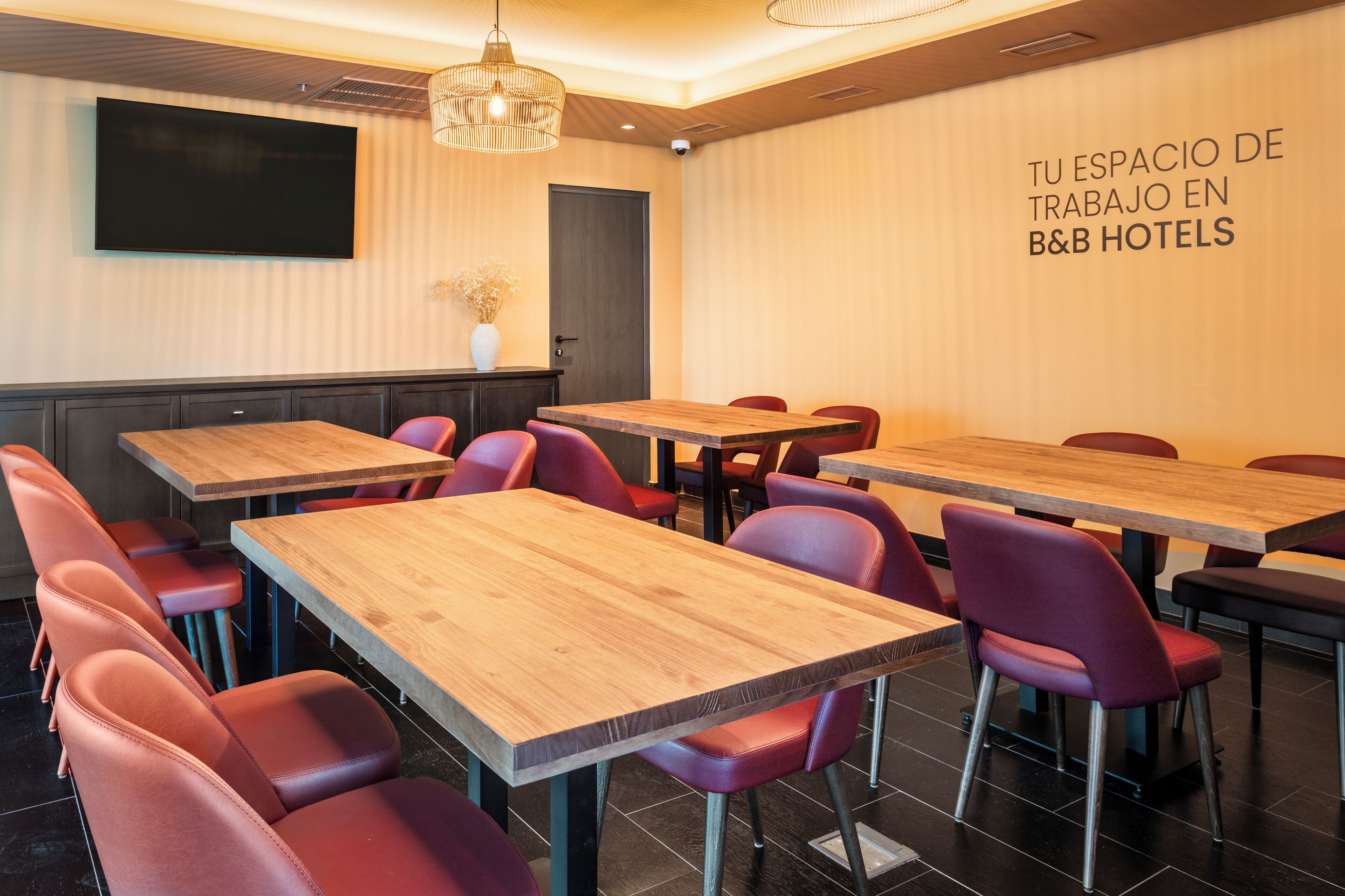 A conference room with multiple tables and chairs, a flat-screen TV mounted on the wall, and a sign indicating it's a workspace at B&B Hotels.