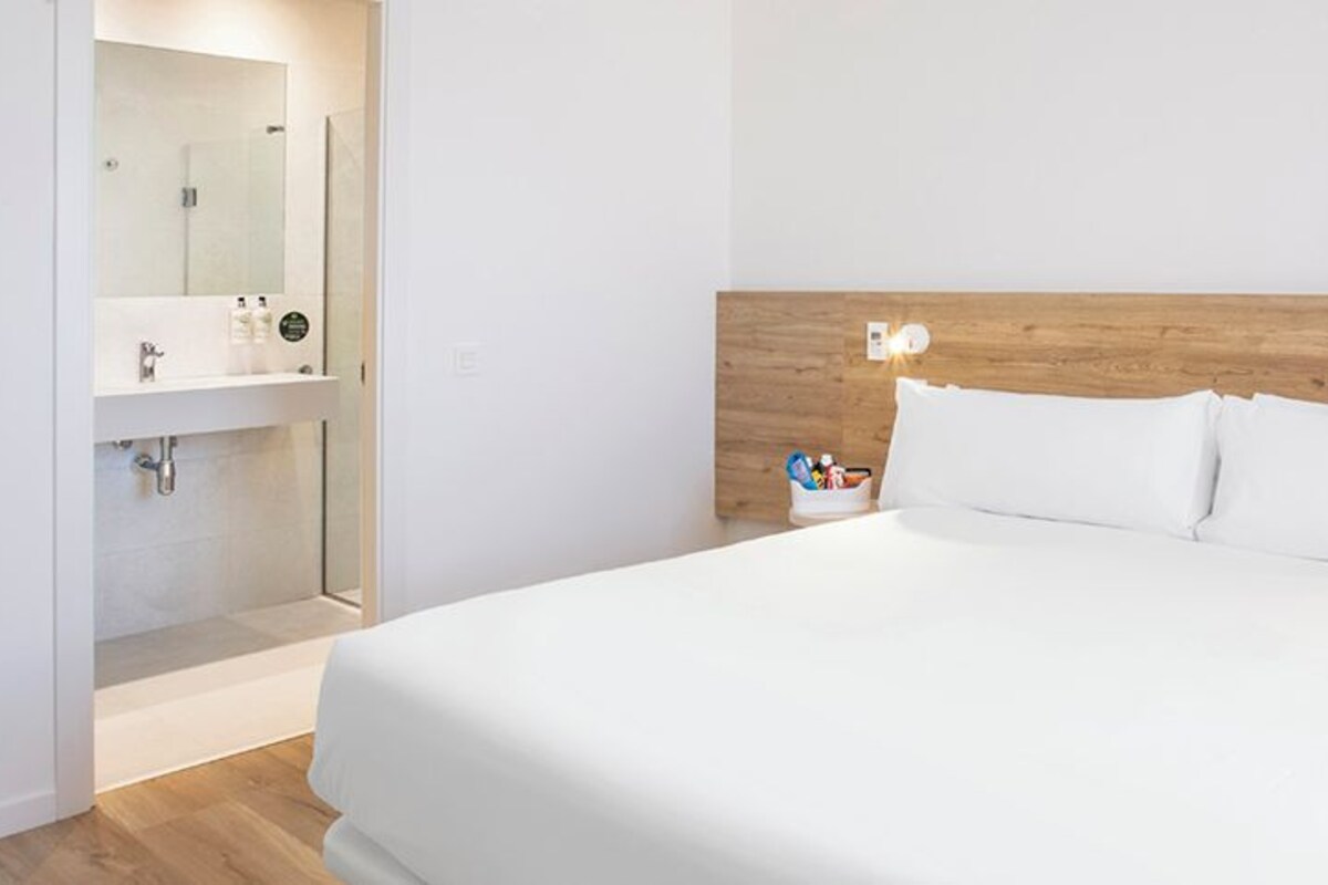 Quarto de hotel com cabeceira de madeira, cama branca, mesa de cabeceira com abajur e banheiro com espelho e pia.