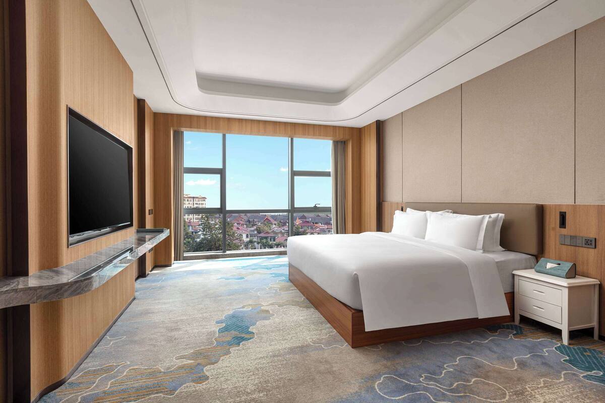 Ein modernes Hotelzimmer mit einem großen Bett, einem Flachbildfernseher, einem Nachttisch und Blick auf eine Stadtlandschaft durch große Fenster.