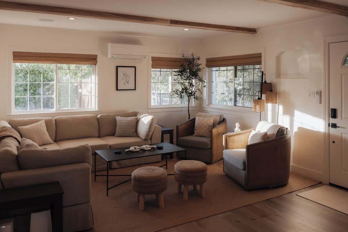 A living room with a sectional sofa, a coffee table, and two ottomans. There are windows with blinds, a potted plant, and a lamp.