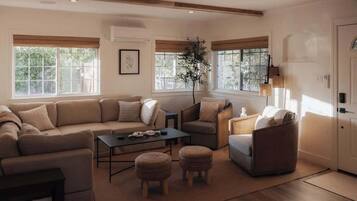 A living room with a sectional sofa, a coffee table, and two ottomans. There are windows with blinds, a potted plant, and a lamp.
