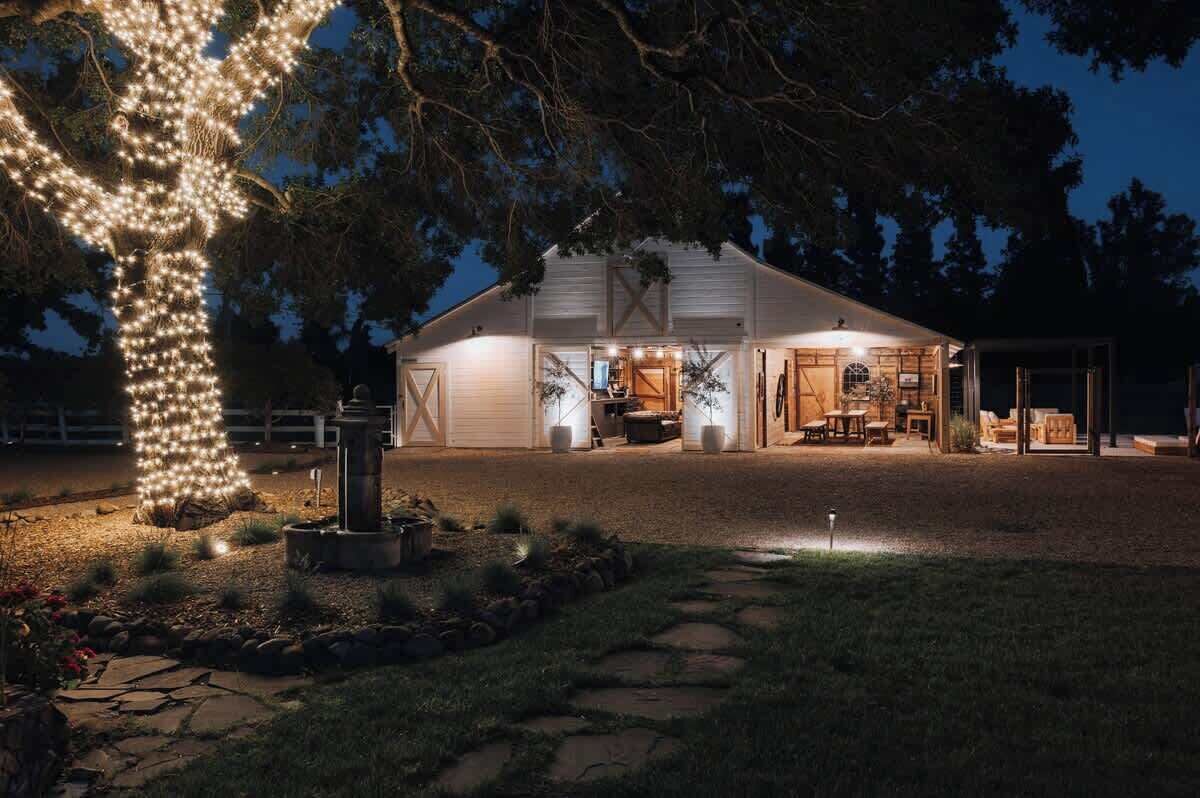 Un bâtiment blanc de style grange, doté d’un patio couvert, d’un chemin de pierre menant à l’entrée et d’un arbre décoré de guirlandes lumineuses.